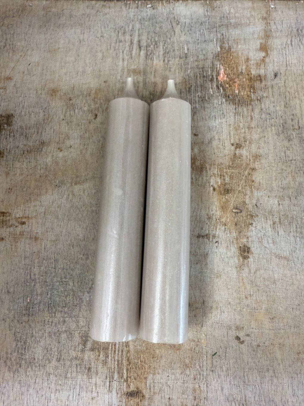 Twee taupekleurige dinerkaarsen naast elkaar, liggend op een houten oppervlak met een verweerde structuur.