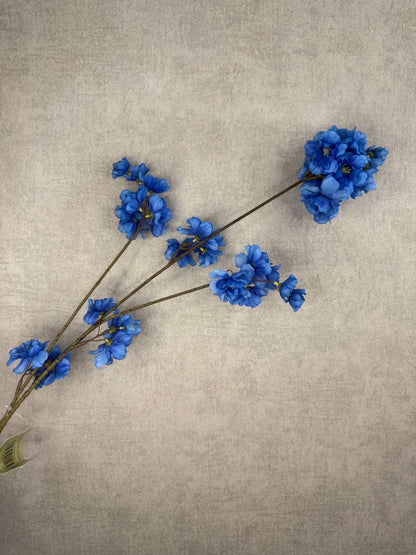 Zijden bloesemtak met helderblauwe bloemen en een fijne vertakking tegen een neutrale achtergrond.