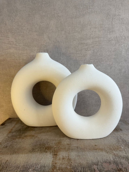 Twee matwitte keramieken donutvazen in Japandi-stijl met ronde openingen, staand op een houten ondergrond tegen een neutrale muur.