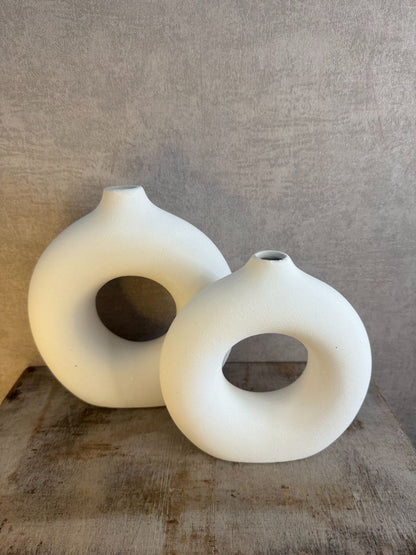 Twee matwitte keramieken donutvazen in Japandi-stijl met ronde openingen, staand op een houten ondergrond tegen een neutrale muur.