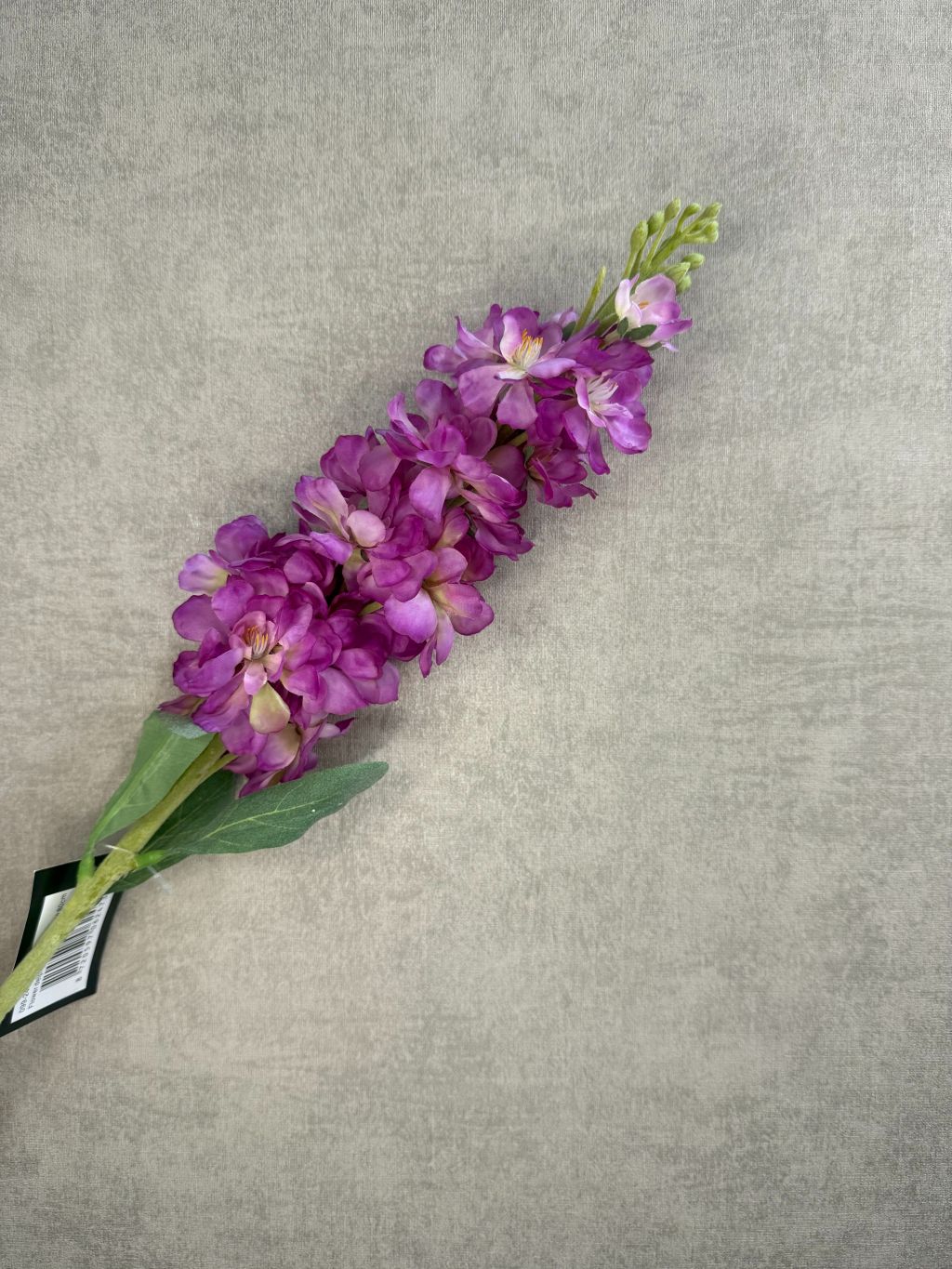 Kunstbloem Delphinium in helder paars met groene bladeren en lange steel, liggend op een lichtgrijze ondergrond.