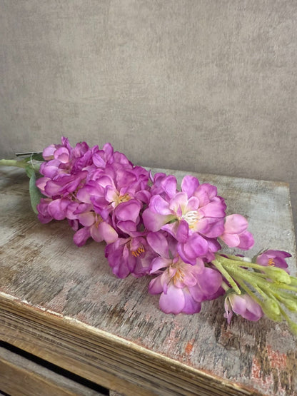 Kunstbloem Delphinium in helder paars met groene bladeren en lange steel, liggend op een lichtgrijze ondergrond.