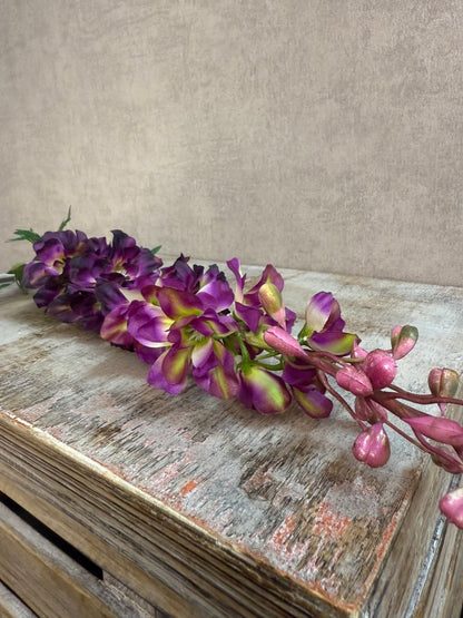 Kunstbloem Delphinium Paars melange met paarse bloemen en groene steel, liggend op een grijze ondergrond.