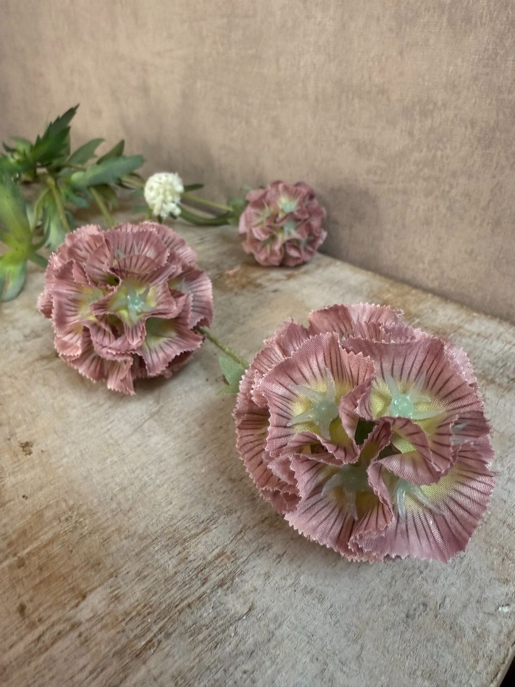 Kunsttak met drie oudroze Scabiosa bloemen en groene bladeren, liggend op een grijze ondergrond met fijne structuur.
