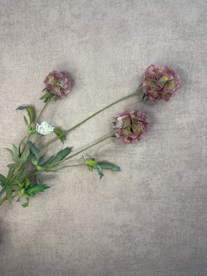 Kunsttak met drie oudroze Scabiosa bloemen en groene bladeren, liggend op een grijze ondergrond met fijne structuur.