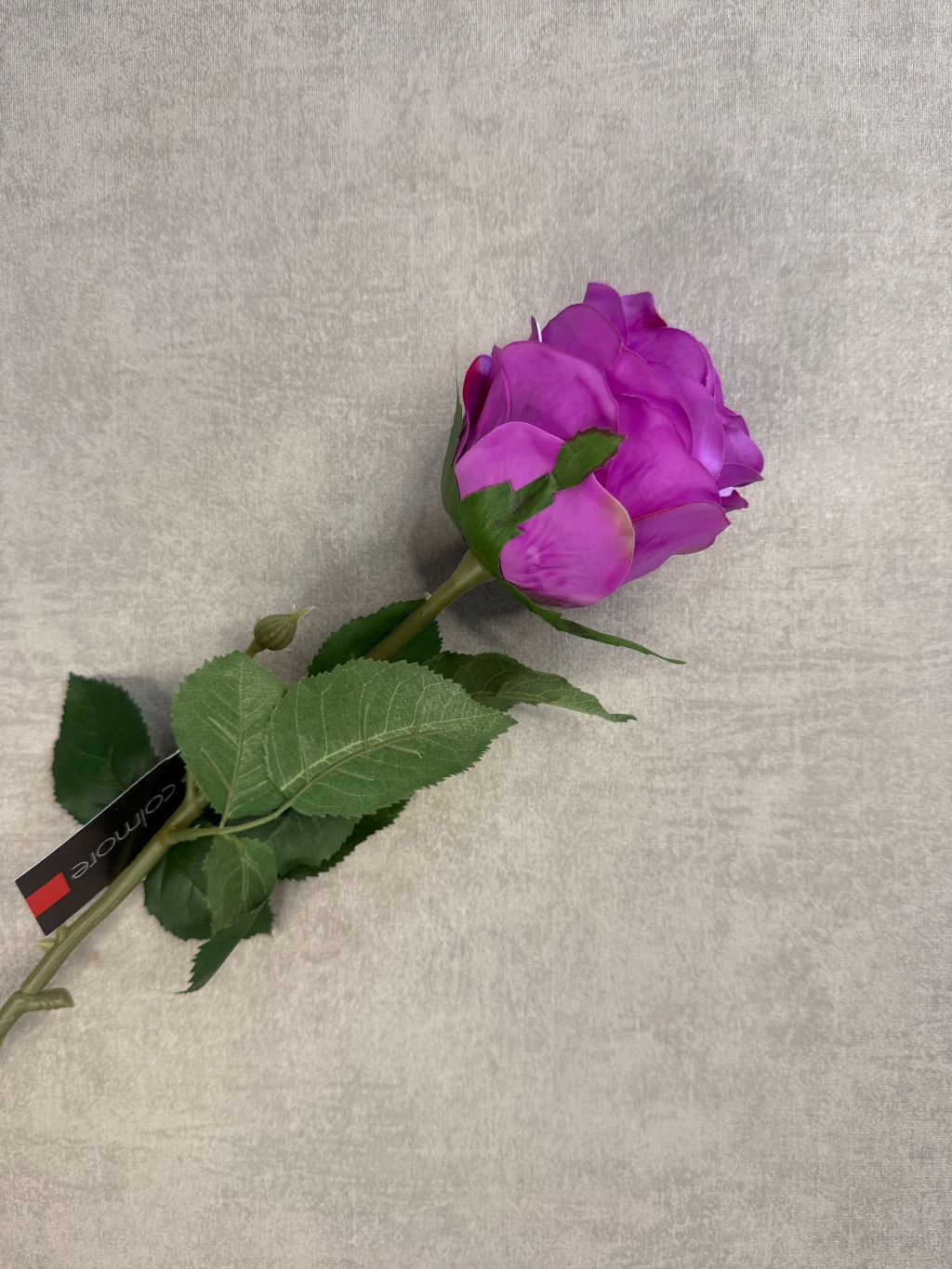 Kunstroos in fel paars met groene bladeren en label van Colmore, liggend op een lichtgrijze ondergrond met fijne structuur.
