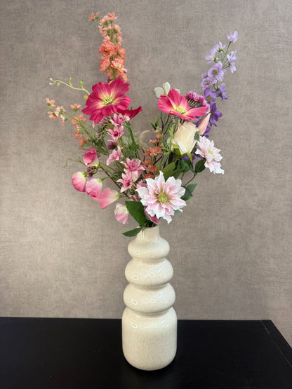 Kunstboeket in bonte kleuren met roze, lila en perzikbloemen in crèmekleurige vaas tegen grijze achtergrond.