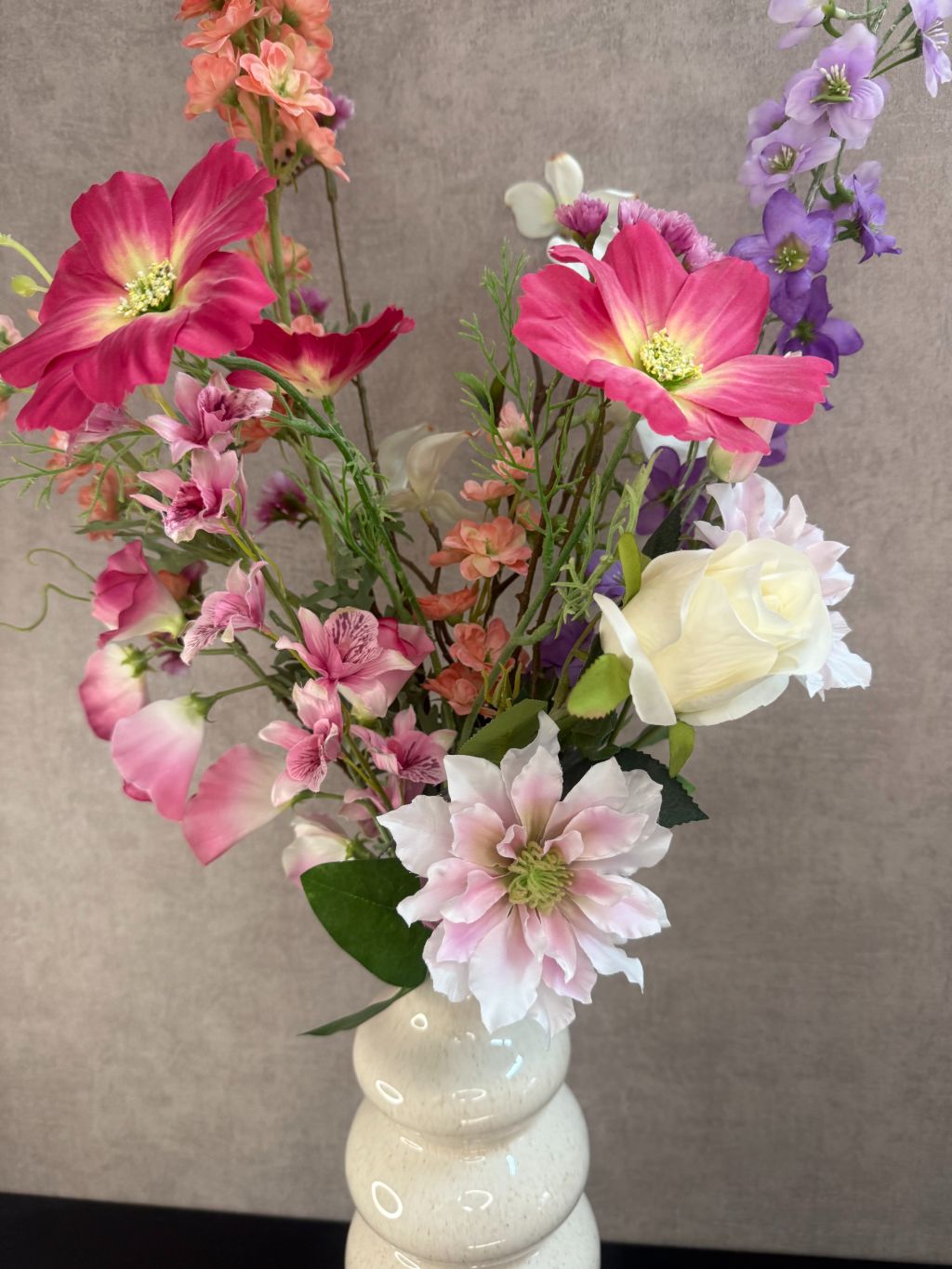Kunstboeket in bonte kleuren met roze, lila en perzikbloemen in crèmekleurige vaas tegen grijze achtergrond.