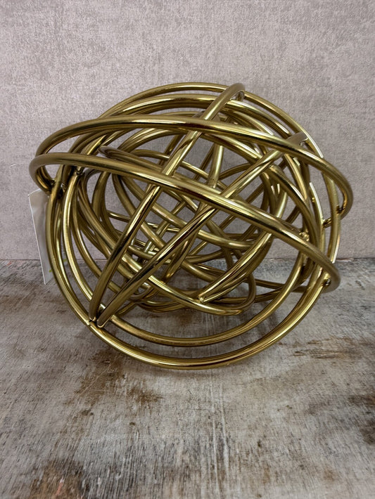 Gouden metalen bolornament met open gekruist lijnenspel op een houten ondergrond.