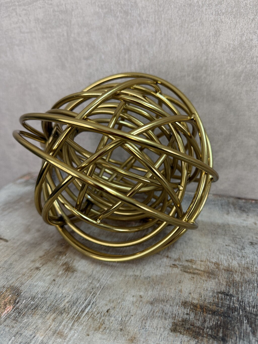 Gouden metalen bolornament met open gekruist lijnenspel op een houten ondergrond.