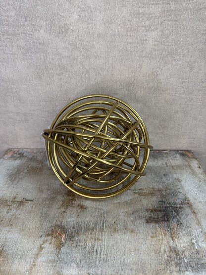 Gouden metalen bolornament met open gekruist lijnenspel op een houten ondergrond.
