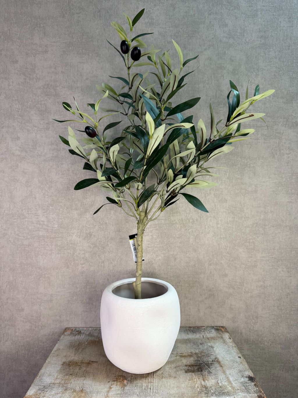 Kunst olijfplant met groene bladeren en zwarte olijven in een witte pot op een houten ondergrond.