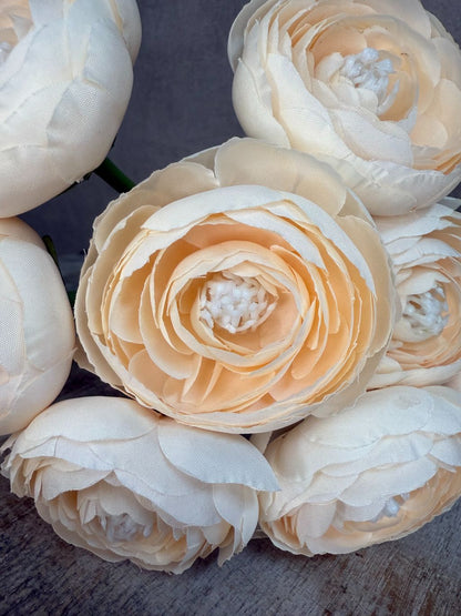 Zijaanzicht van meerdere kunstbloemen ranonkel in champagnekleur met lange steel, liggend op een houten ondergrond.