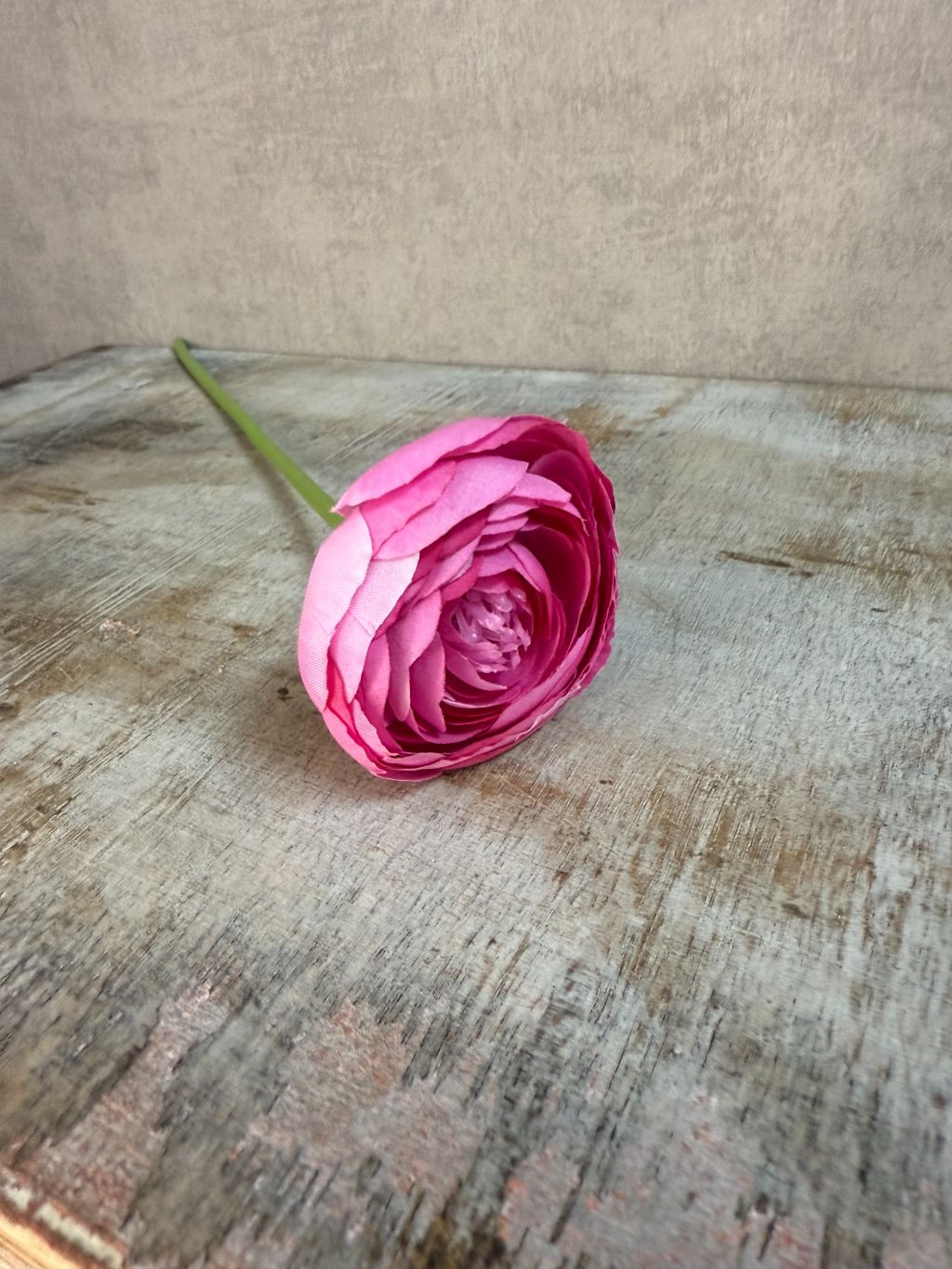 Kunstbloem ranonkel in roze tint met realistische bloemstructuur en groene steel, liggend op een houten oppervlak.