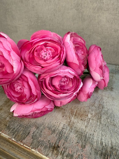 Meerdere Kunstbloemen ranonkel in roze tint met realistische bloemstructuur en groene steel, liggend op een houten oppervlak.