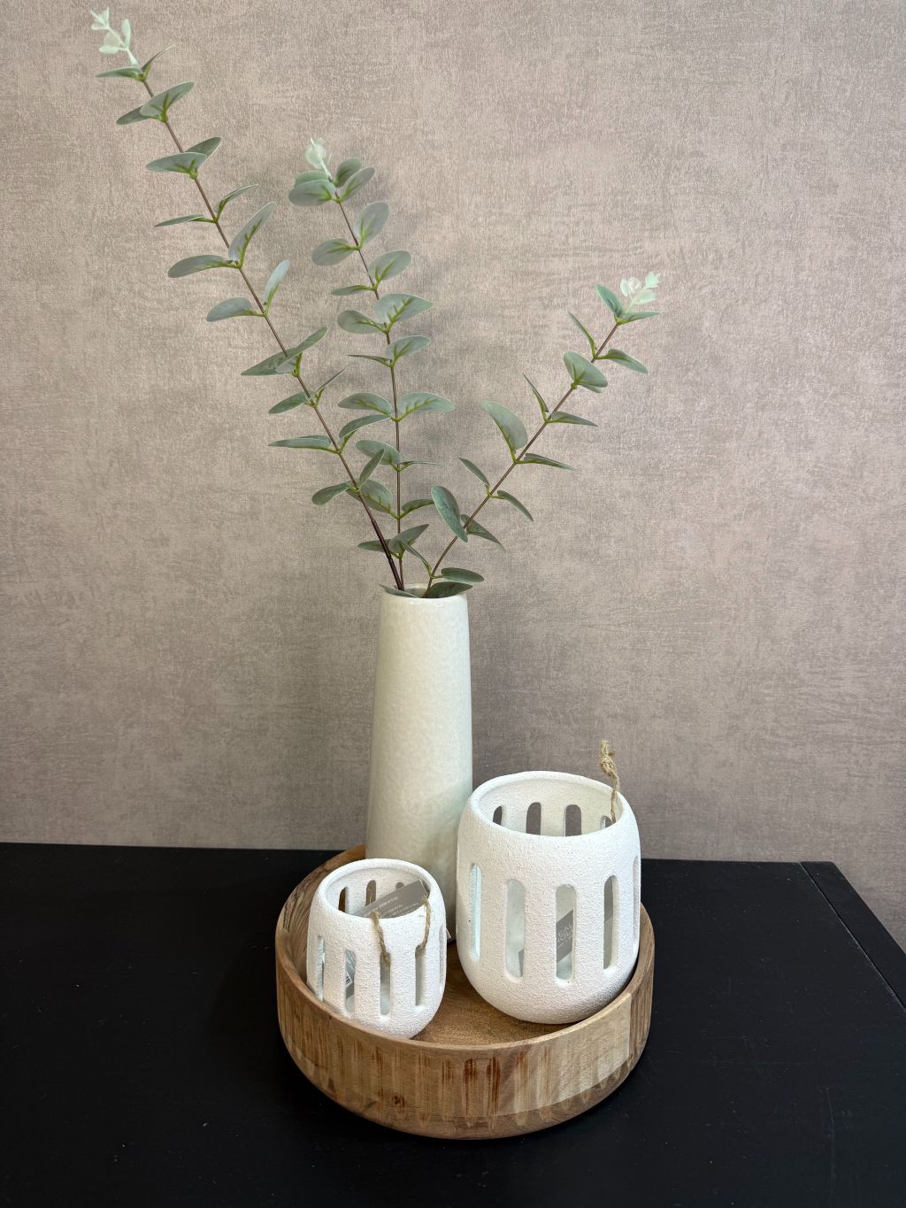 Rond houten dienblad met crème vaas met groene takken en twee crème waxinelichthouders op een zwarte tafel.