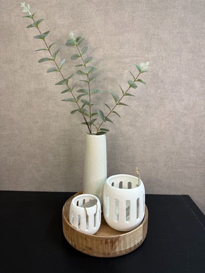 Rond houten dienblad met crème vaas met groene takken en twee crème waxinelichthouders op een zwarte tafel.