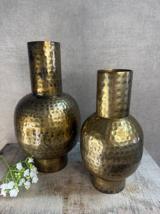 Twee metalen vazen in antiek goud met hamerslagstructuur, staand op houten ondergrond tegen een grijze muur. Eén bloemtak op de voorgrond.