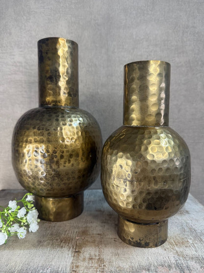 Twee metalen vazen in antiek goud met hamerslagstructuur, staand op houten ondergrond tegen een grijze muur. Eén bloemtak op de voorgrond.