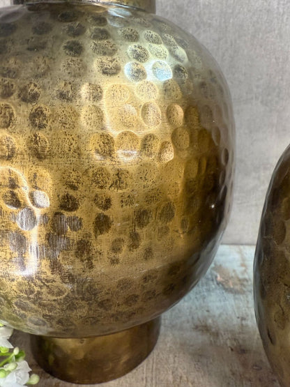 Twee metalen vazen in antiek goud met hamerslagstructuur, staand op houten ondergrond tegen een grijze muur. Eén bloemtak op de voorgrond.