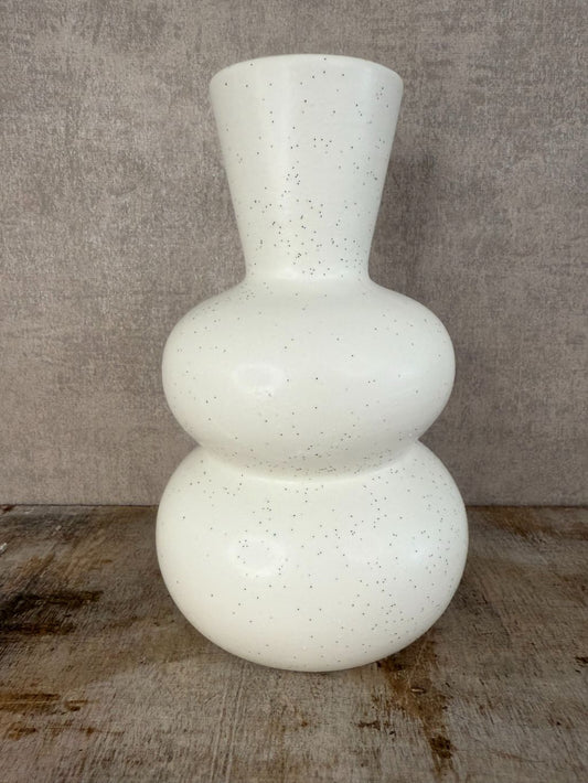 Witte keramieken vaas met zwarte spikkels en bolvormig design, staand op een houten oppervlak tegen een grijze achtergrond.