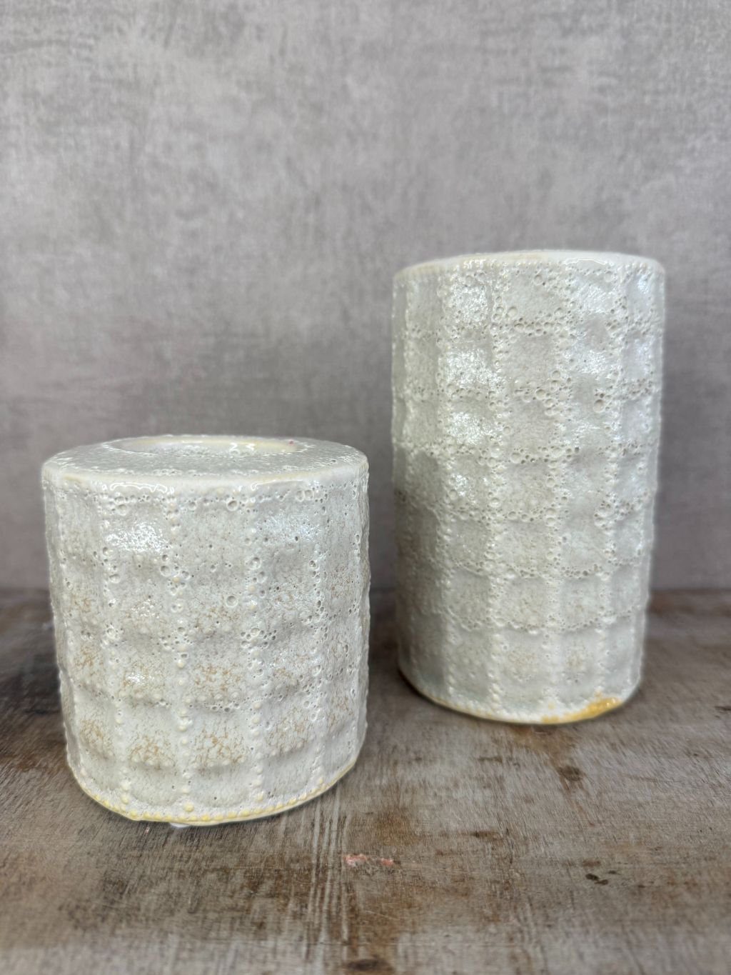 Twee keramieken waxinelichthouders in beige/grijs met lichte structuur en glans, geplaatst op een houten ondergrond.