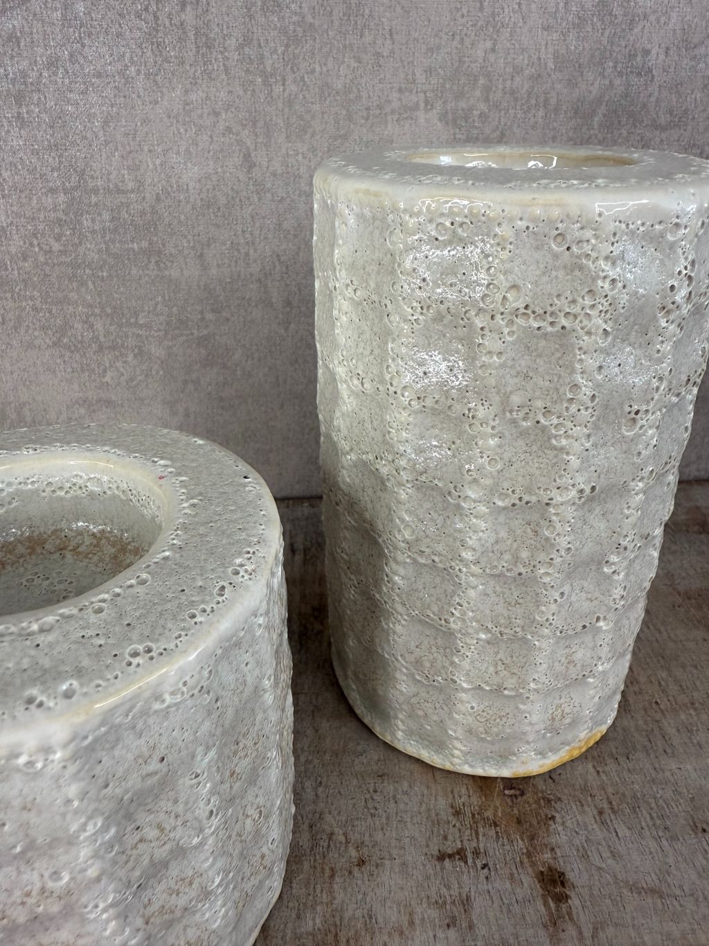 Twee keramieken waxinelichthouders in beige/grijs met lichte structuur en glans, geplaatst op een houten ondergrond.